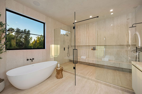 Modern luxurious bathroom with a freestanding tub, large glass shower, and a view of trees through a window.