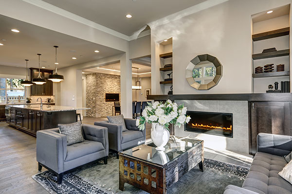 Chic living room filled with built-in cabinets flanking round mirror atop grey tile fireplace, tufted sofa facing two armchairs and window wall overlooking  lush outdoors. Northwest, USA