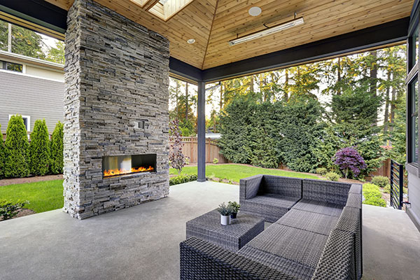 New modern home features a backyard with covered patio accented with stone fireplace, vaulted ceiling with skylights and furnished with gray wicker sofa placed on concrete floor. Northwest, USA